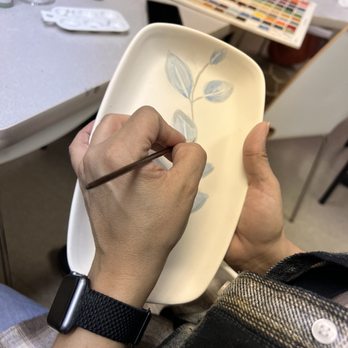 The Painted Giraffe - A person is painting a leaf pattern on a ceramic dish in an art studio, with various art supplies and color charts visible in the background. - Art Classes Near Me - Westmont, Illinois