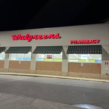 Walgreens - Nighttime exterior of Walgreens pharmacy, showing the building's facade with illuminated red Walgreens sign and pharmacy sign. The storefront has multiple windows with green awnings, and the parking lot in front is visible with some pavement markings and a small puddle. The scene is dark except for the signage and interior lighting visible through the windows. - Cosmetics & Beauty Supply Near Me - Severna Park, Maryland