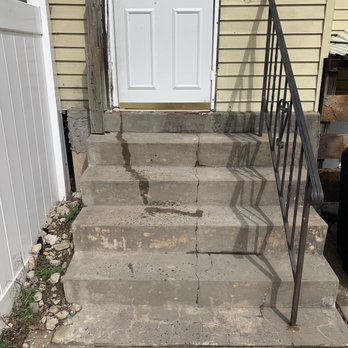 Fr Contracting - Cracks in the steps - Concrete stairs with five steps, showing multiple cracks and water stains, lead up to a door with a white frame and a small window. The stairs are adjacent to a beige siding wall, with a black metal railing on the right side. The left side of the stairs has a wooden post and some debris and rocks at the base. The concrete surface appears aged and weathered, with visible cracks and discoloration. - Roofing Near Me
