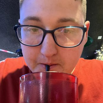 Players Pub - Me drink a Dr pepper - A young man with short, light brown hair and black glasses is holding a large red cup with a straw, drinking from it. He is wearing a red shirt and is seated indoors, with a dark wall and some decorative lights visible in the background. - Sports Bars Near Me - Tucson, Arizona