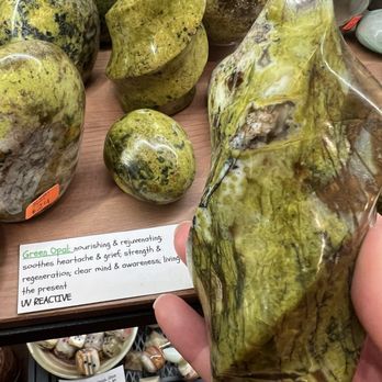 Dimitridon Studios - A large, polished mineral specimen with a predominantly green color and yellowish-brown streaks is held in a person's hand. The background shows other polished stones and mineral specimens on display, with a small handwritten sign describing the mineral as 'Green Opal' and its properties. The setting appears to be a mineral and gemstone shop with various rocks, fossils, and crystals available for sale. - Jewelry Near Me - Alexandria, Kentucky