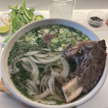 Phoxotic - The Butcher's Choice Pho - A bowl of pho featuring beef, sliced onions, and chopped green onions, with a clear broth. Side herbs, lime, and condiments are visible nearby. - Vietnamese Near Me - Washington, DC, District of Columbia