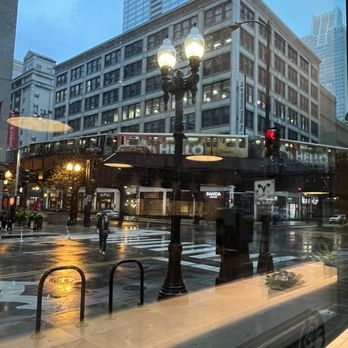 Two Zero Three - The L - City street scene viewed through a window with reflections, featuring buildings, a train, pedestrians, and streetlights on a rainy day. - Coffee & Tea Near Me - Chicago, Illinois