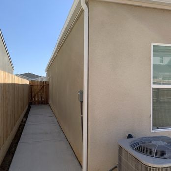 ProPatio by Weber Construction - A concrete walkway is positioned alongside a beige stucco house, with a window visible on the right side. An air conditioning unit is situated at the corner of the house, with a pipe connecting it to the building. A wooden fence lines the left side of the walkway, leading to a gate at the end. The sky above is clear and blue. - General Contractors Near Me