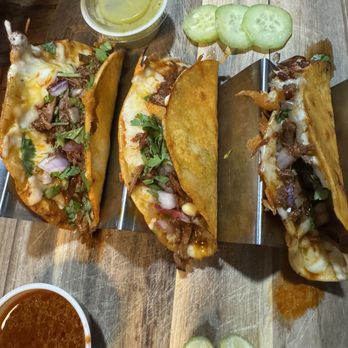 Daddy Shark - Birria tacos - Three birria tacos filled with beef, melted cheese, and topped with cilantro and onions are arranged on a wooden serving board. The tacos are partially open, revealing the filling inside. Two small cups of sauce, one red and one green, are placed nearby, along with slices of lime and cucumber. The presentation suggests a Mexican cuisine setting, emphasizing the traditional dish of birria tacos with garnishes and dipping sauces. - Seafood Near Me - Clinton, Mississippi