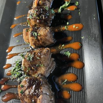 Flavorture - "Deep fried" salmon bites - Four pieces of glazed meat are arranged on a black plate, topped with sauce and garnished with chopped herbs, with additional sauce drizzled around. - Bars Near Me - Washington, DC, District of Columbia