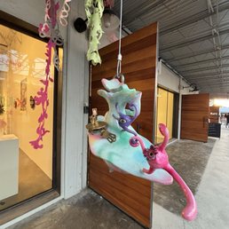 A hanging sculpture of a whimsical, colorful cat with exaggerated features is positioned outside a storefront. The sculpture is pink with additional colors and details, and it appears to be made of a lightweight material. It is suspended by a string and is located near a wooden panel wall and a glass window of the shop. The scene is set on a sidewalk with a metal roof overhead.