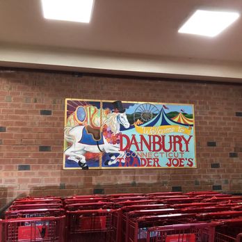 Trader Joe's - The image shows a mural painted on a brick wall inside a store or shopping area. The mural includes a carousel horse wearing a top hat and a floral necklace, with circus tents and a Ferris wheel in the background. The text on the mural reads 'Welcome to Danbury, Connecticut, Trader Joe's.' Below the mural, there are red shopping carts stacked together. The ceiling has bright rectangular lights illuminating the space. - Grocery Near Me - Danbury, Connecticut