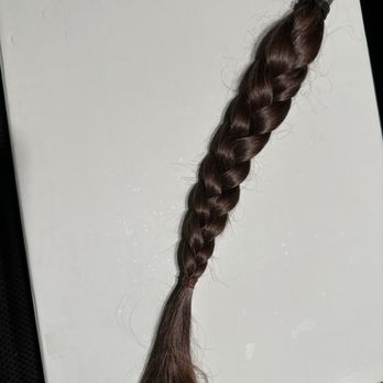 Dolce Salon - 12-inches of 'virgin hair' ready for donation to  Locks of Love - A single braid of brown hair is positioned on a white surface, with the hair tied at the end. The braid appears to be freshly cut, with some loose strands at the end. The surface is plain and unadorned, providing a clear view of the hair's texture and color. - Hair Salons Near Me - Austin, Texas