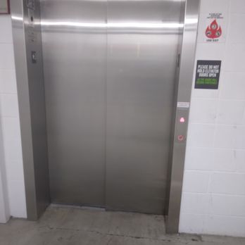 Extra Space Storage - The image shows a stainless steel elevator with closed doors, positioned against a white tiled wall. To the right of the elevator, there are safety and operational signs, including instructions not to hold the doors open and a fire safety notice. The floor appears to be concrete with visible cracks. The setting suggests an indoor public or commercial building environment. - Self Storage Near Me - Berwyn, Illinois