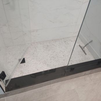 Belmore Glass & Mirror - The image shows a shower enclosure with clear glass walls and a black metal frame. The interior walls are covered with white marble tiles with subtle gray veining. The shower floor is made of small, white pebble tiles. A black square drain is visible on the floor, and a handheld showerhead is mounted on the wall. The enclosure is situated on a gray tiled bathroom floor. - Glass & Mirrors Near Me
