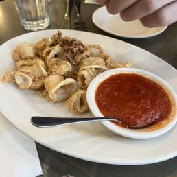 Totally solid calamari - Fried calamari pieces are arranged on a white oval plate, with a small bowl of marinara sauce. A spoon rests on the plate's edge. In the background, a glass of water with a lemon wedge and a person's hand holding a lemon wedge are visible. The setting appears to be a restaurant or diner table.