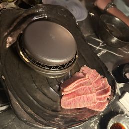 Wagyu - Thin slices of marbled beef are arranged on a black grill plate at a restaurant, with various dishes and utensils visible around.