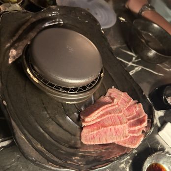 Sakaba - A5 Wagyu Hot Rock - Thin slices of marbled beef are arranged on a black grill plate at a restaurant, with various dishes and utensils visible around. - Sushi Bars Near Me - Avon, Colorado