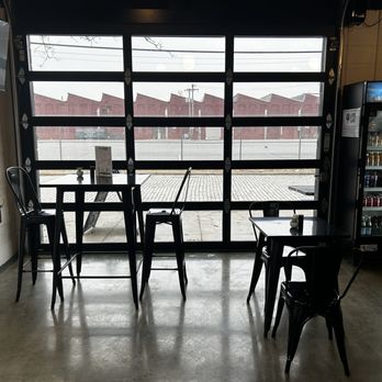 LIFE Cafe - Seating with a tv. - The image shows the interior of LIFE Cafe with a concrete floor, several black metal tables and chairs, and a large garage-style window that provides a view of an outdoor area with brick buildings. A vending machine stocked with drinks is positioned on the right side of the image. The space appears to be minimally decorated with a focus on functional seating and natural light from the window. - Coffee & Tea Near Me - Pawtucket, Rhode Island