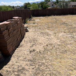Before 1 - An outdoor backyard area with a dirt and sparse grass ground, enclosed by a wooden fence, and a stack of bricks on the left side.