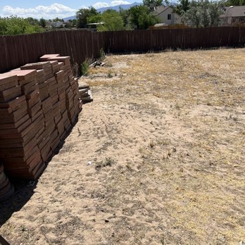 Alpha1 Sprinklers - Before 1 - An outdoor backyard area with a dirt and sparse grass ground, enclosed by a wooden fence, and a stack of bricks on the left side. - Irrigation Near Me - Albuquerque, New Mexico