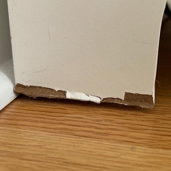 BZ Movers - The image depicts a close-up of the bottom corner of a white door or cabinet, where the cardboard backing is torn and chipped, exposing the inner material. The object is positioned on a wooden floor, with the damage clearly visible at the junction of the door and the floor surface. - Movers Near Me - Long Grove, Illinois