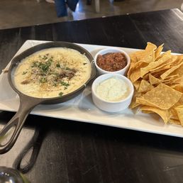 Happy hour tavern chips. The cheese dip is quite good! - A rectangular white serving plate contains a small cast iron skillet of melted cheese dip garnished with herbs, two small bowls of salsa and sour cream, and a large portion of tortilla chips. The plate is placed on a dark wooden table in a restaurant setting, with some blurred background elements visible.