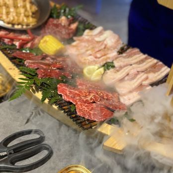 Y Space BBQ - Display of the Dragon boat - A selection of raw meats, including beef, chicken, and seafood, along with vegetables and lemon slices, are laid out on a grill with smoke rising. The presentation suggests a dining experience involving grilling or BBQ, with additional dishes and utensils visible nearby. - Barbeque Near Me - Flushing, New York