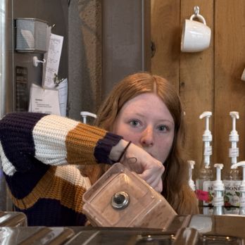 Foundry Coffeehouse - A woman with red hair and a striped sweater is sitting at a cafe counter, holding a blender, with a wooden wall and coffee mugs in the background. - Coffee & Tea Near Me - Lenexa, Kansas