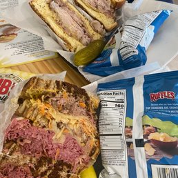 The image shows two sandwiches with sliced meats, cheese, and vegetables, placed on a tray. One sandwich appears to contain corned beef or pastrami, while the other has a different type of deli meat, with visible shredded cheese and vegetables. A pickle and a bag of Ruffles potato chips are also on the tray. The setting suggests a casual dining environment, likely at a deli or sandwich shop.