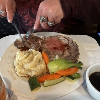 Vallemar Station - A person, wearing rings on their fingers, is cutting into a medium-rare steak on a white plate. The meal includes mashed potatoes, cooked carrots, zucchini, and other vegetables. There are small bowls of gravy and sauce on the table, and the setting appears to be a restaurant with a white tablecloth. - Steakhouses Near Me - Pacifica, California