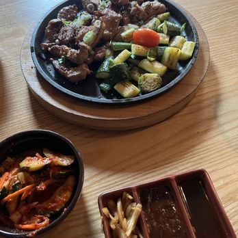 Char'd - Hand-Cut Grilled Pork Ssam - A hot cast iron skillet contains grilled pork pieces with chopped green onions, served with zucchini, cucumber, and a cherry tomato. Side dishes include kimchi and dipping sauces. - Korean Near Me - Milwaukee, Wisconsin