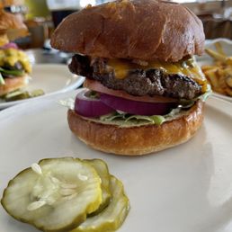 Green Chile Cheeseburger - A cheeseburger with a beef patty, cheddar cheese, tomato, onion, and lettuce on a bun, served with pickles on a white plate.