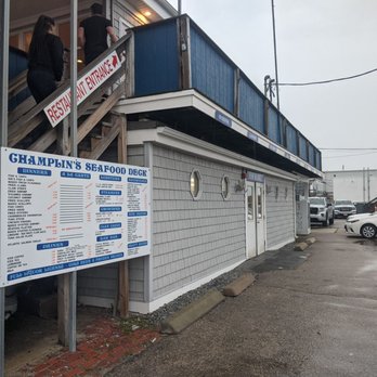 Champlin's Seafood - Entrance - The photo depicts the exterior of Champlin's Seafood Deck, a waterfront seafood establishment. A large menu sign is positioned near the building, listing various seafood dishes, drinks, and other offerings. The building has a staircase leading to an upper deck, with a couple of people visible on the stairs. The parking lot in front contains multiple parked cars, and the sky appears overcast. - Seafood Near Me - Narragansett, Rhode Island