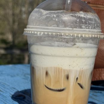 Command Coffee - Iced  Cold Brew Coffee - A cold coffee drink with a layer of foam on top features a smiley face drawn in the foam, with two eyes and a curved mouth. The beverage is in a clear plastic cup with a dome lid and a straw, set on a blue wooden surface outdoors. The background shows blurred trees and natural scenery, indicating an outdoor setting. - Coffee & Tea Near Me - Indianapolis, Indiana