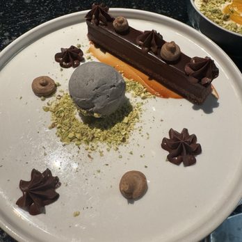 ilili DC - ilili Candy Bar - A dessert plate contains a long rectangular chocolate pastry with decorative chocolate dollops on top, a scoop of gray ice cream on crushed green pistachio, and small dollops of chocolate cream. The plate is white and set on a dark surface, with a bowl of additional dessert visible in the background. - Lebanese Near Me - Washington, DC, District of Columbia