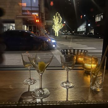Double Fun - Bar - The image shows a nighttime view from inside a bar, with three cocktail glasses on a wooden table, two of which contain lemon slices. There are candles in holders on the table, casting a warm glow. Outside the window, city street lights, a lit tree, and a building with large windows are visible. A car is passing by on the street, and the scene suggests an urban setting with a lively atmosphere. - Cocktail Bars Near Me - Chicago, Illinois