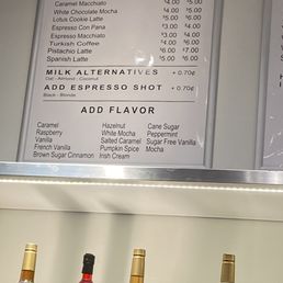 Menu - A menu board titled 'Coffee & More' lists coffee drinks such as latte, Americano, flat white, cappuccino, and others, with prices for regular and large sizes. Additional options include milk alternatives, espresso shots, and flavor add-ons. Below the menu, bottles of flavored syrups are visible, indicating customization options for drinks.