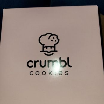 Crumbl - Naperville - Peanut butter chocolate chip - A white box from Crumbl Cookies displays a black cartoon dog logo above the brand name. The logo depicts a simple, stylized dog face with a rounded head, floppy ears, and a collar. The brand name 'crumbl' is written in lowercase letters, with 'cookies' underneath in smaller font. The box appears to be held in a person's lap, with dark clothing visible in the background. - Bakeries Near Me - Naperville, Illinois