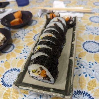 Rolly Kimbab - Crab salad kimbap - A rectangular ceramic plate contains sliced kimbap rolls with seaweed wrapping, revealing fillings such as vegetables and possibly seafood or meat. The plate is on a table with a patterned cloth featuring yellow and blue designs. In the background, there are small dishes with side items, a blue cup, chopsticks, and other dining utensils, indicating a meal setting at a restaurant or home. - Korean Near Me - Flushing, New York