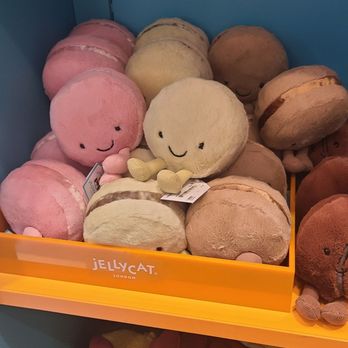 Jellycat - Plush toys resembling macarons with smiling faces in pink, beige, and brown are arranged on a shelf at Jellycat in Chicago, IL. The toys have soft, fuzzy textures and small limbs, with some featuring additional details like tiny arms. The display includes a variety of colors and sizes, with other plush toys visible underneath, including slippers and a bowl-shaped plush item. The shelf is part of a retail display for Jellycat plush products. - Toy Stores Near Me - Chicago, Illinois