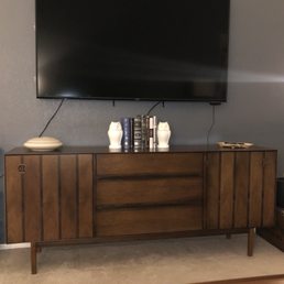Our refinished credenza - A wooden sideboard featuring a mid-century modern design with vertical paneling and tapered legs. On top, there are two white owl-shaped vases, a collection of books, a ceramic dish, and a decorative object. A large flat-screen TV is mounted on the wall above the sideboard, with visible cables hanging down. The setting appears to be a living room with a neutral wall color and a beige carpeted floor.