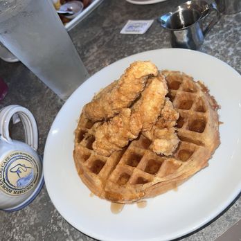 Another Broken Egg Cafe - A plate with fried chicken tenders on top of a waffle, drizzled with syrup, on a restaurant table with drinks and bread. - Breakfast & Brunch Near Me - Fishers, Indiana