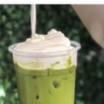 TeaJito - Matcha boba's good - A cold matcha milk tea with black tapioca pearls at the bottom is presented in a clear plastic cup. The drink has a vibrant green color and is topped with whipped cream. The background is blurred with green foliage, and part of a white sign with black text is visible at the top. - Bubble Tea Near Me - Miami, Florida