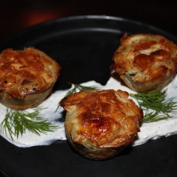 Nerina - The image shows three baked Greek-style appetizers, possibly stuffed eggplants or similar, arranged on a black plate. They are topped with a golden-brown cheese or sauce and placed over a white sauce with herbs, likely dill. The presentation emphasizes the Mediterranean cuisine style, with a focus on traditional ingredients and plating. - Greek Near Me - Brooklyn, New York