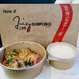 Two takeout containers of dim sum are placed in front of a cardboard box with restaurant branding. One container holds a mixture of vegetables and seafood, while the other contains a white dipping sauce. The cardboard box behind features the restaurant's name, 'Juicy Dumplings,' with additional text and design elements. The setting appears to be a dining or takeout area, with the focus on the food packaging and branding.