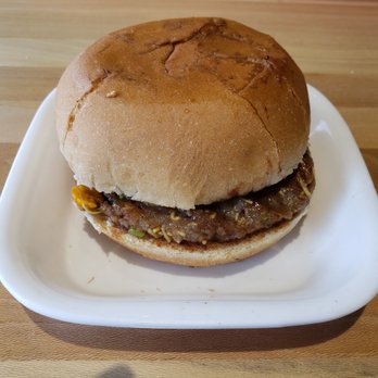 Neehee's Hanover Park, Illinois - Dabeli - A burger with a thick, textured patty inside a round bun, served on a white plate on a wooden table. - Indian Near Me - Hanover Park, Illinois