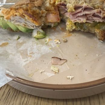 Meta Cafe - Turkey on the left, ham on the right. Fantastic flavors and delicious bread! - A sandwich with layers of turkey, ham, and avocado is cut in half and placed on a paper-lined plate. The bread appears toasted, and the fillings are visible inside. The plate is on a wooden table, with a small dish in the background. - Sandwiches Near Me - Lambertville, New Jersey