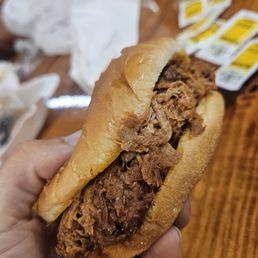 Pulled Pork - A close-up of a pulled pork sandwich being held in a person's hand. The sandwich contains shredded pork meat inside a soft bun. In the background, there are several barbecue sauce packets on a wooden table, indicating a casual dining setting, likely at a barbecue restaurant or eatery.