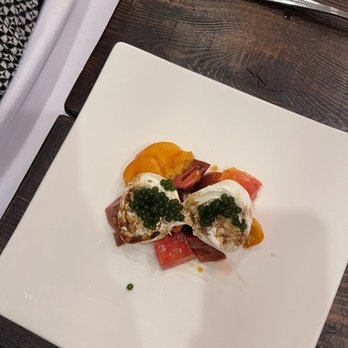 The Twisted Tines - Two poached eggs topped with green caviar are arranged on a white square plate. The eggs are accompanied by diced yellow and red peppers, creating a colorful presentation. The plate is set on a dark wooden table with utensils placed nearby, indicating a fine dining setting. - Personal Chefs Near Me