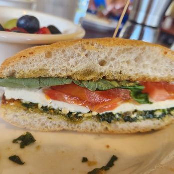 Il Vinaio - Caprese Chiabatta - A close-up of a sandwich featuring fresh greens, sliced tomato, and cheese inside a crusty bread roll. The sandwich is served on a plate with some herbs and crumbs visible. In the background, there are bowls of berries and a person using a smartphone, indicating a casual dining setting. - New American Near Me - Mesa, Arizona