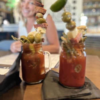 Hash Kitchen - BYO Bloody - Two Bloody Mary drinks with skewers of olives, cheese, bacon, and a pickle, placed on black napkins on a wooden table, with a smiling woman in the background. - New American Near Me - Gilbert, Arizona