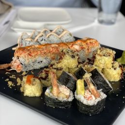 Shrimp tempura, Vegas crunch, volcano II, and spicy California - A black plate features various sushi rolls, including tempura and maki, served with garnishes on a white table.