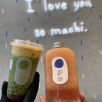 Machi Machi - Red Bean Brûlée Matcha Smoothie, Grapefruit Slush with Grapefruit Jello - A person wearing a black glove holds a green tea drink with tapioca pearls in a clear cup, and another person holds a pinkish beverage in a clear bottle with a silver cap. The background features a wall with a neon sign that reads 'I love you so machi,' surrounded by colorful confetti-like decorations. - Bubble Tea Near Me - Markham, Ontario